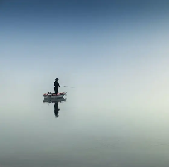 A person stands alone in a boat on a still, misty lake